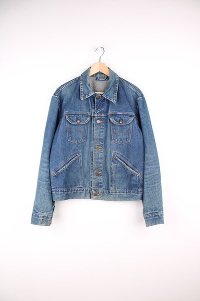 Vintage Wrangler denim trucker jacket with branded button closure, snaps on the cuffs, four front pockets and zig-zag stitch detailing. Small blue and yellow Wrangler logo on left breast pocket. 