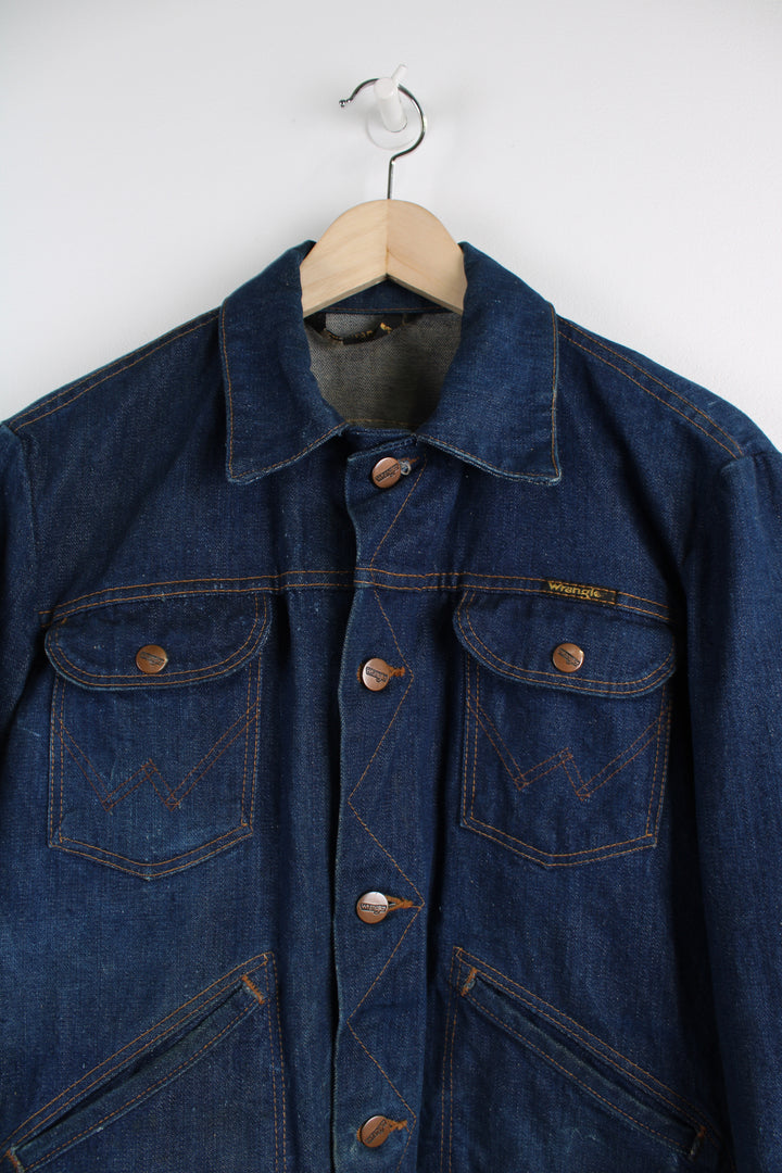 Vintage Wrangler Denim Jacket in a blue colourway with brown cross stitching, button up, multiple pockets, and has logo embroidered on the chest.