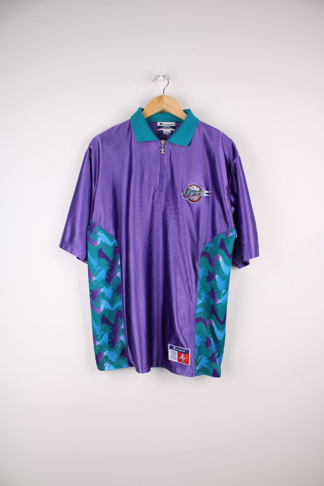 Vintage Champion Utah Jazz polo shirt style jersey in a purple colourway with a turquoise collar, and zip closure. The shirt has patterned side panels and the Utah Jazz logo embroidered on the front. There is also a Champion NBA authenticity patch sewn on the hem.