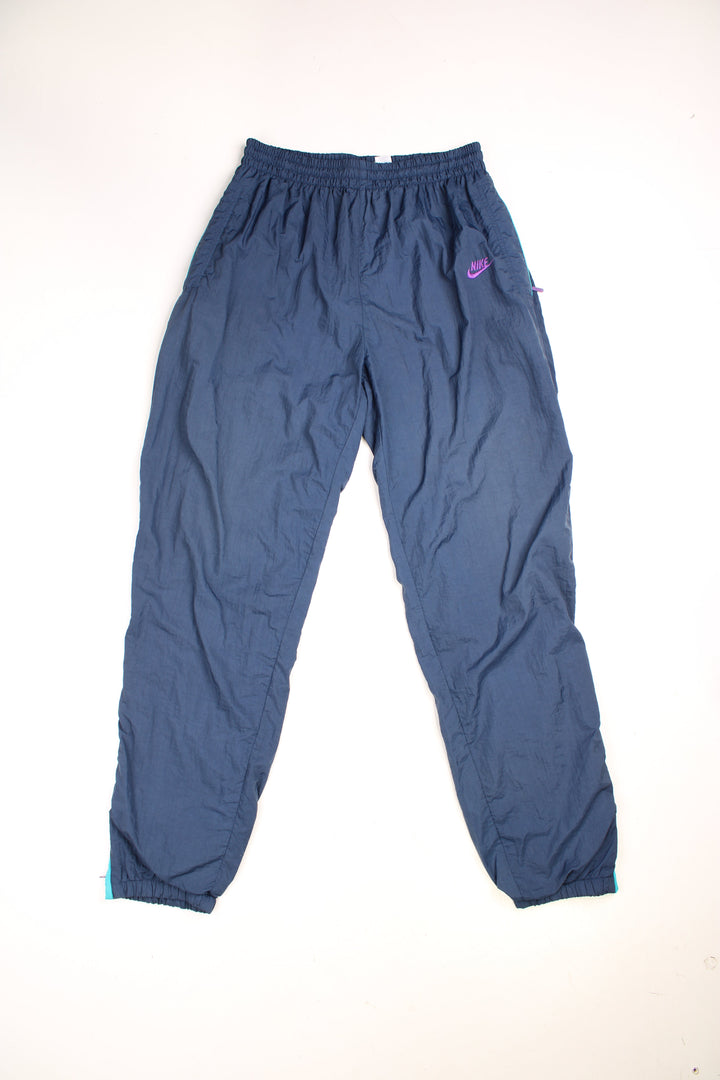 Vintage Nike Shell Tracksuit Bottoms in a blue colourway, has an elasticated waist with drawstring, zip up side pockets, and the logo embroidered in purple on the front.