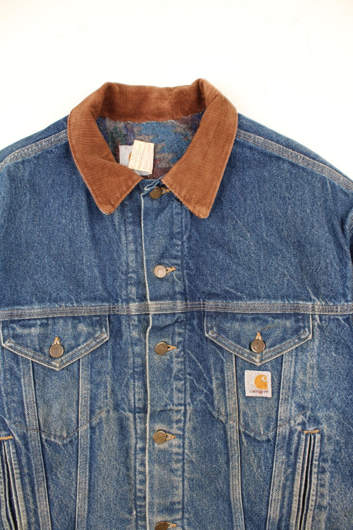 Vintage Carhartt Denim Jacket in a blue colourway with a brown corduroy collar. Buttons up and has multiple pockets, insulated with a blanket lining, and has the logo embroidered on the front.