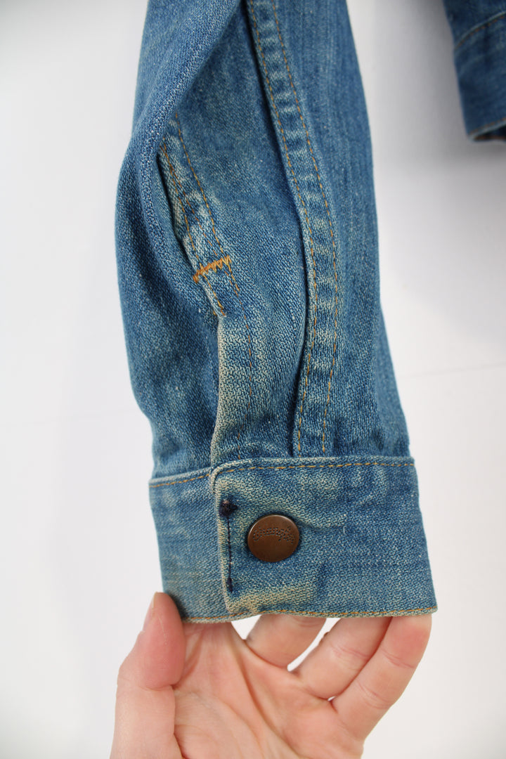 Vintage Wrangler Denim Jacket in a blue colourway with brown cross stitching throughout, button up, multiple pockets, and has logo embroidered on the chest.