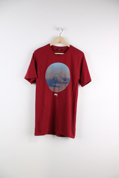 Stussy Graphic T-Shirt in a red, blue and brown colourway, has the logo printed in the centre underneath mountain graphic design.