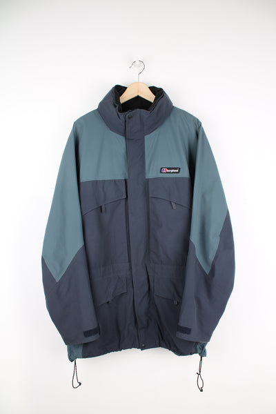 Berghaus Jacket in a grey and blue colourway, zip up, multiple pockets, hidden hood, waterproof and has logo embroidered on the front. 
