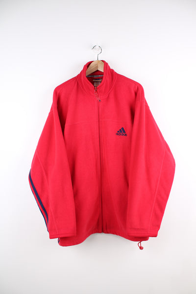 Adidas Fleece in a red colourway, navy blue iconic three stripes going down the sides, zip up, side pockets, and has the logo embroidered on the front.