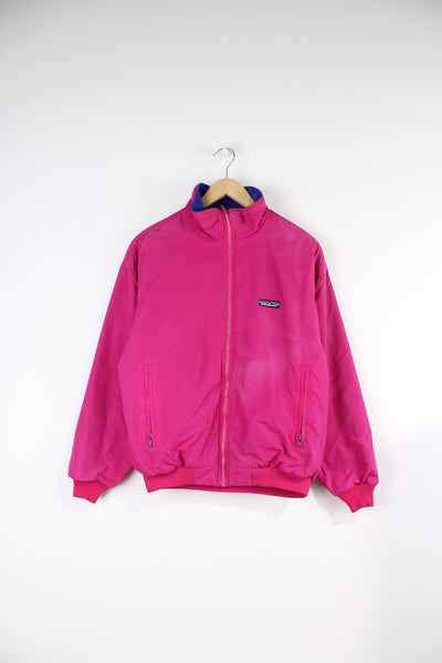 Patagonia Jacket in a pink colourway and has a purple fleece lining, zip up, pockets, and has logo embroidered on the front. 