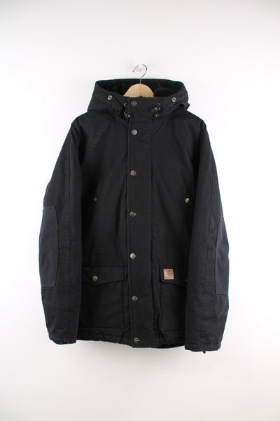 Carhartt Parka Jacket in a black colourway, zip up, multiple pockets, hooded with a faux fur lining and insulated with a quilted lining, and has the logo embroidered on the front.