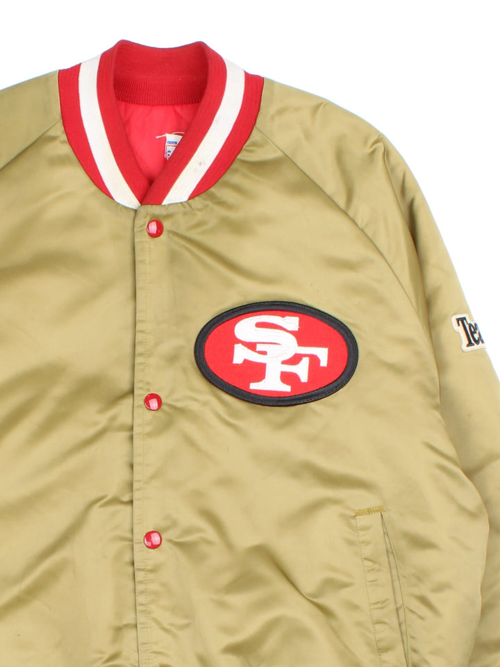 Vintage San Francisco 49ers NFL Chalk Line Varsity Jacket in a gold tan colourway. Buttons up and has side pockets, insulated with a quilted lining, and has the team logo and spell out embroidered on the front and back.