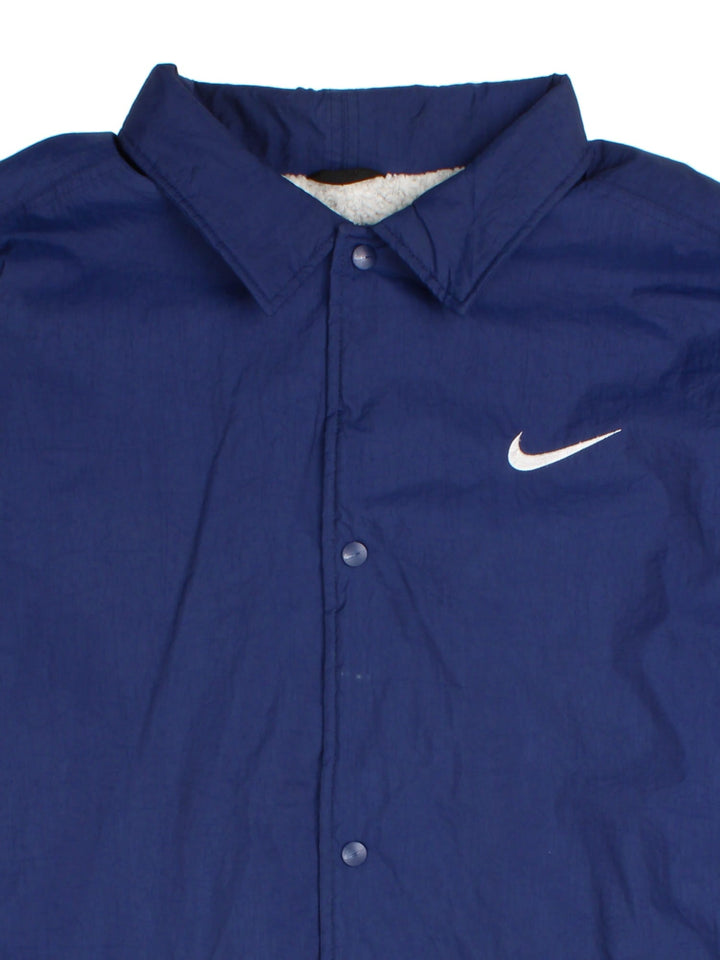 Nike Coach Jacket in a blue colourway. Fleece lining with full button up front, double zip pocket with small classic nike swoosh. Large Nike logo across the back.