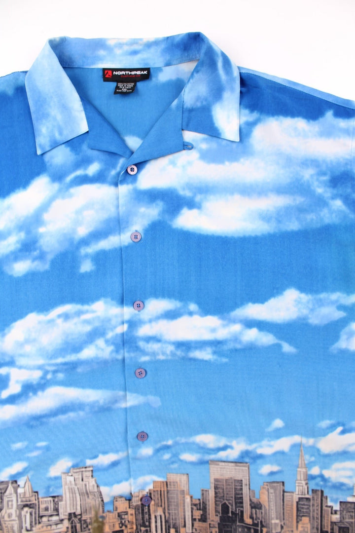 Vintage Northpeak shirt in blue with print of the New York City skyline against a blue sky and clouds.