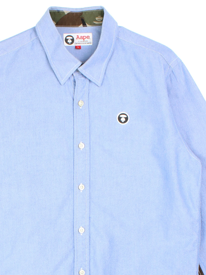 A Bathing Ape By Bape Oxford Shirt in a blue colourway and has camo panelling by on the sleeves, button up with the logo embroidered on the front.