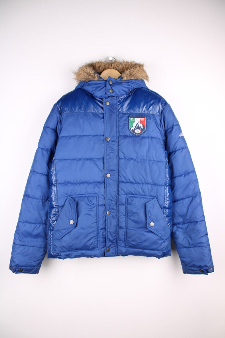Y2K Ellesse Coat in a blue colourway, the coat has a faux fur trim hood, is filled with a polyester filling and has two hand warmer pockets on the front panels. There is an Ellesse Ski patch sewn onto the front and Ellesse embroidered on the sleeve. 