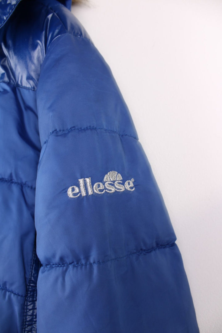 Y2K Ellesse Coat in a blue colourway, the coat has a faux fur trim hood, is filled with a polyester filling and has two hand warmer pockets on the front panels. There is an Ellesse Ski patch sewn onto the front and Ellesse embroidered on the sleeve. 