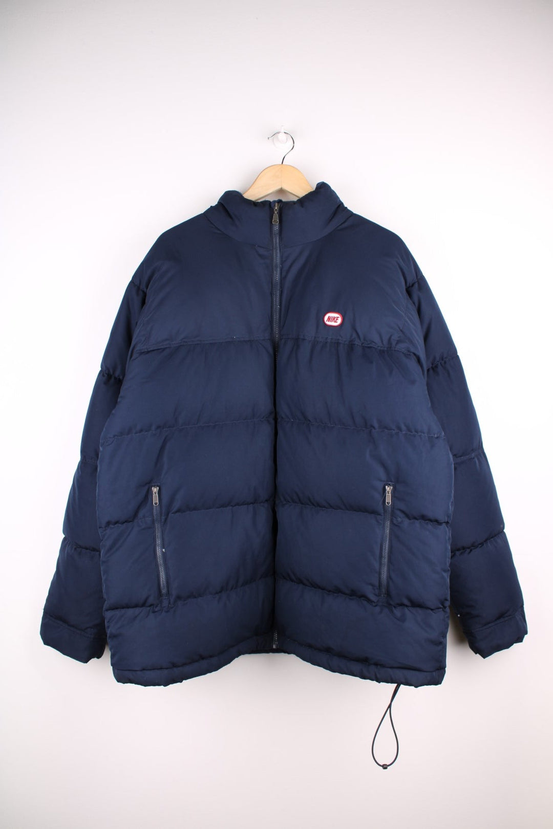 Vintage Nike Y2K era Puffer Coat in a navy blue colourway. The coat is filled with duck feather, has two zip pockets on the front panels and a zip away hood. There is also a small Nike logo stitched on the front. 