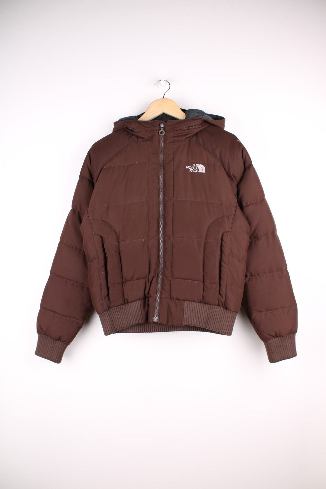 The North Face Puffer Coat in brown. The jacket is filled with duck down feathers, has two pockets on the front panels and a puffer hood. There is an interior pocket, and elasticated cuffs and hem. 