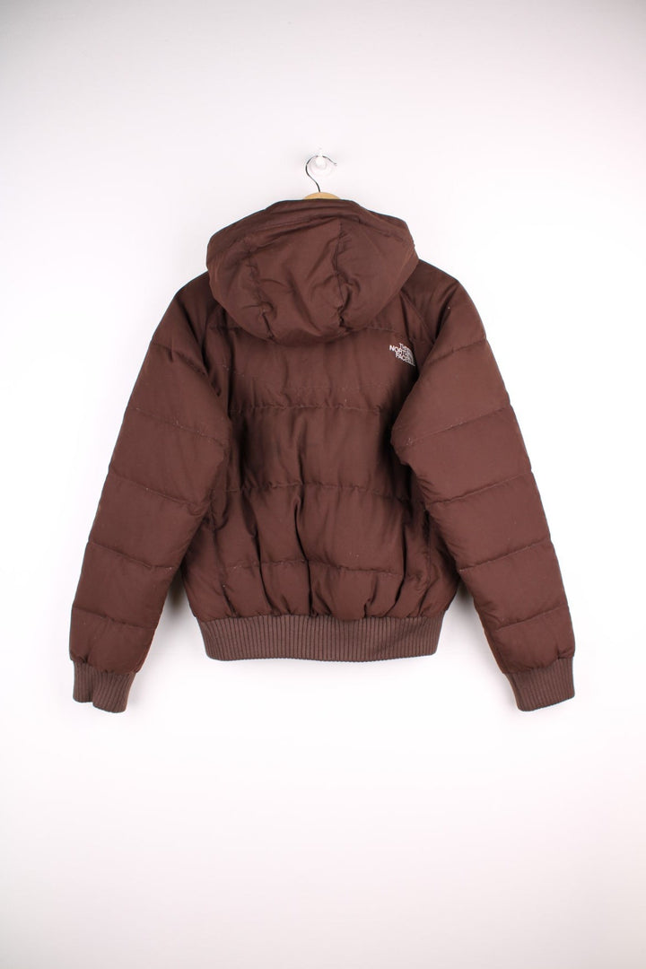 The North Face Puffer jacket in brown. The jacket is filled with duck down feathers, has two pockets on the front panels and a puffer hood. There is an interior pocket, and elasticated cuffs and hem. 