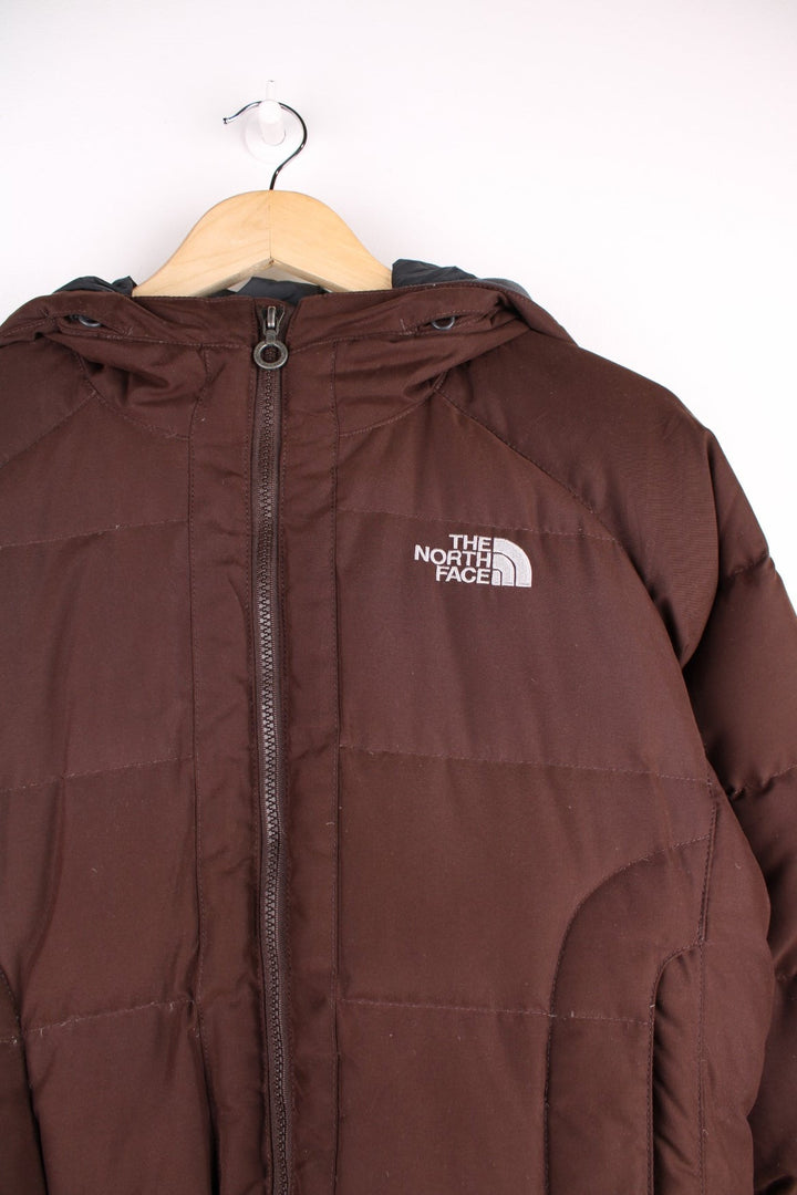 The North Face Puffer jacket in brown. The jacket is filled with duck down feathers, has two pockets on the front panels and a puffer hood. There is an interior pocket, and elasticated cuffs and hem. 