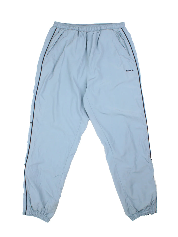 Reebok Tracksuit Bottoms in a blue colourway with stripes going down the sides, pockets, elasticated waist with drawstring, and the logo embroidered on the front and down the sides.
