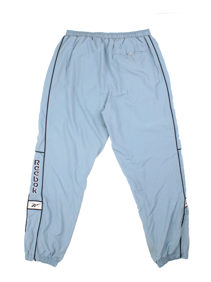 Reebok Tracksuit Bottoms in a blue colourway with stripes going down the sides, pockets, elasticated waist with drawstring, and the logo embroidered on the front and down the sides.