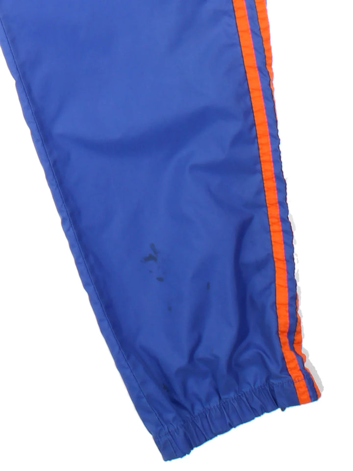 80's Adidas Shell Pants in a blue colourway with the iconic three stripes in orange down the sides, has side pockets, elasticated waist with drawstring, and the logo embroidered on the front.