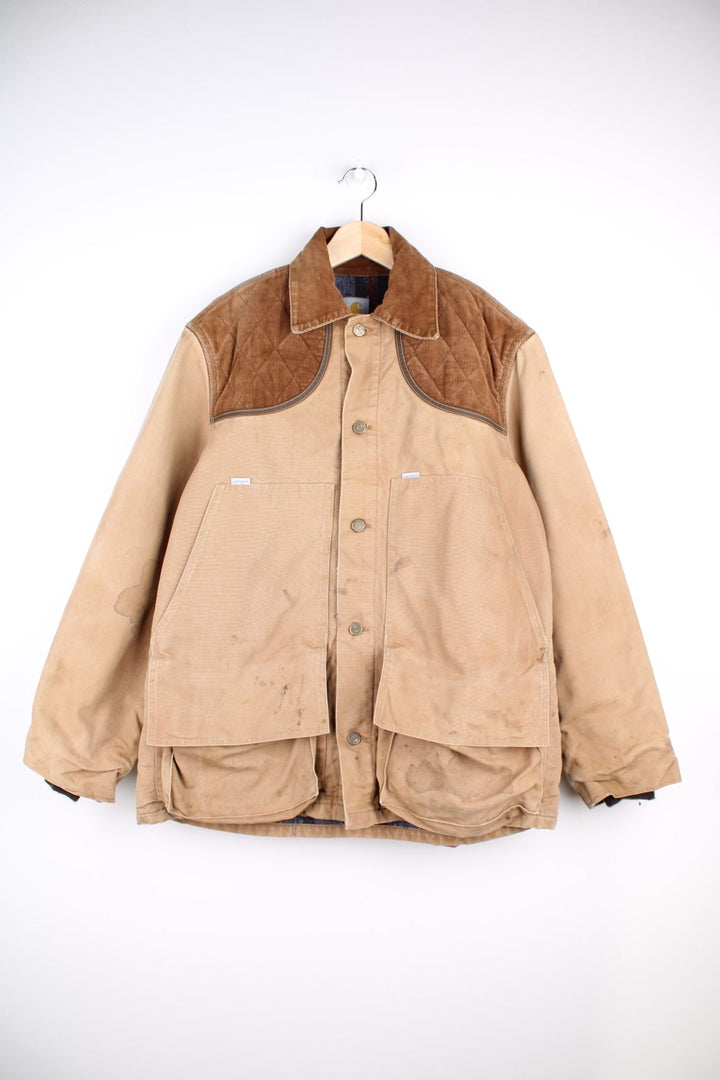 Carhartt Hunting Jacket in a tan browwn colourway with cordurory collar and shoulder pads. Buttons up and has multiple utility pockets including a big detachable pouch pocket on the back, insulated with a blanket lining, and has the logos embroidered on the front.