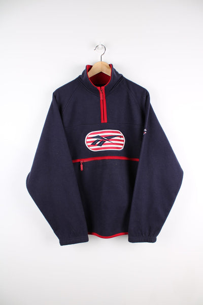 Blue pullover Reebok fleece with large central embroidered logo on the chest as well as a smaller logo on the arm. Features red detailing, a quarter zip and a zip through pocket.
